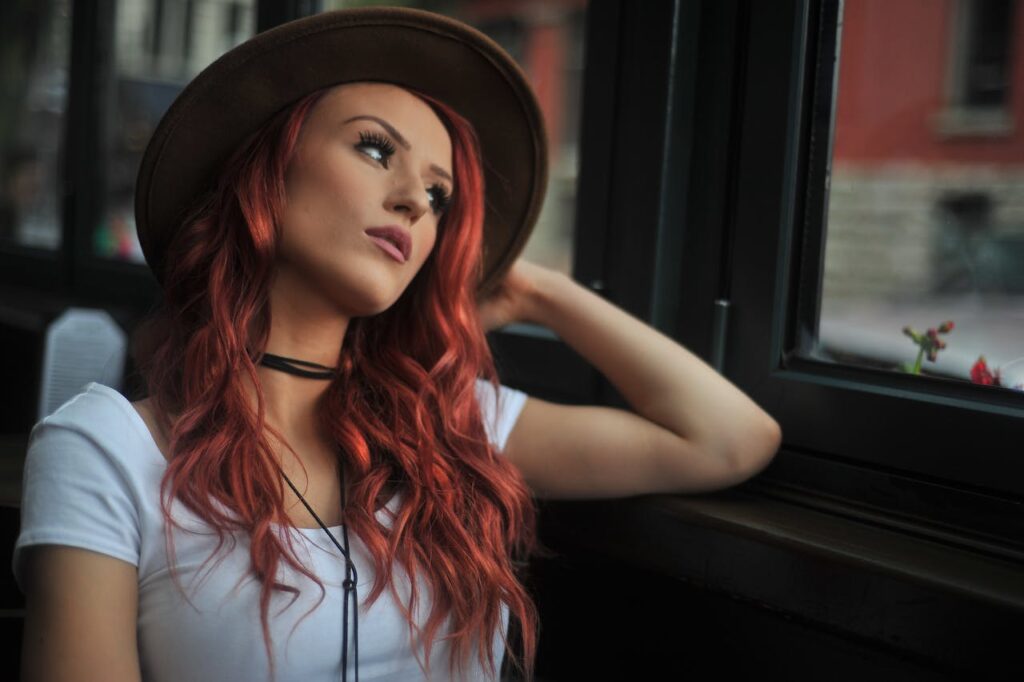 Dívka v klobouku s nabarvenými vlasy hledící z okna.