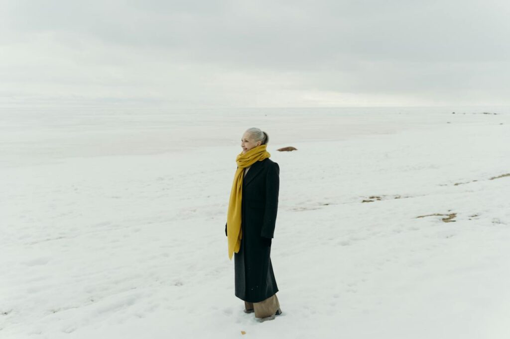 Starší žena v kabátu v zasněžené krajině.