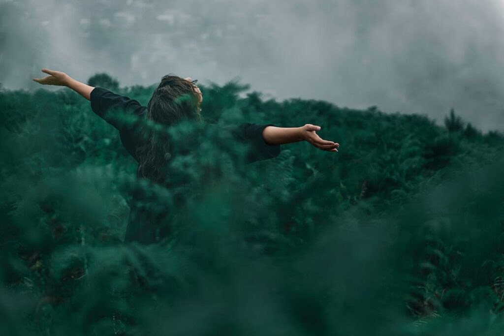 Žena s rozpaženýma rukama obklopená zeleným kapradím.