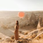 Žena stojící v poušti na pozadí broskvového letícího balónu.