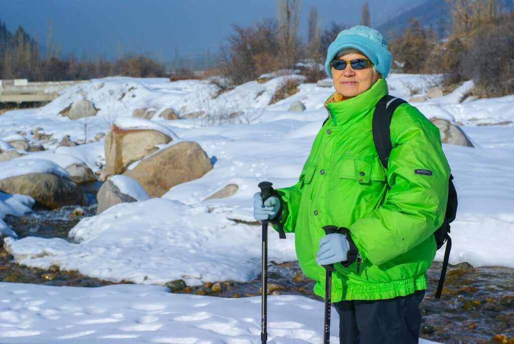 Starší žena v bundě a čepici s turistickými hůlkami v zasněžené krajině.