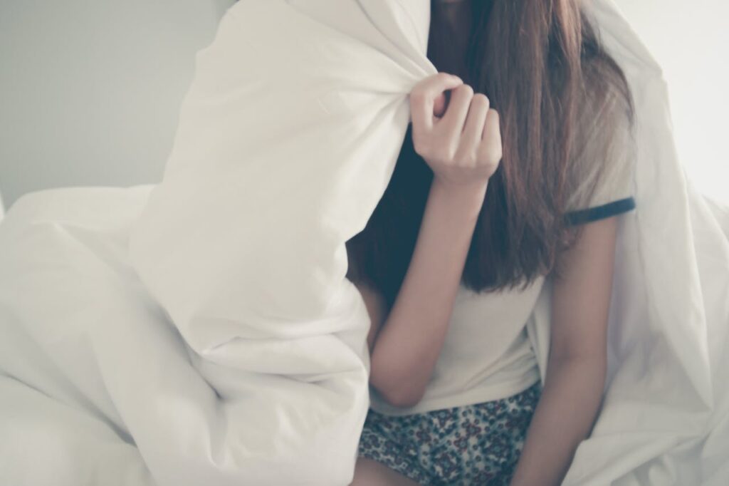 Dívka sedící na posteli přikrytá peřinou.