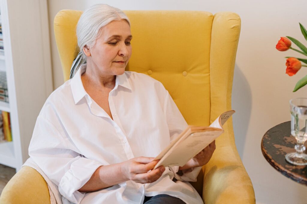 Žena s bílými staženými vlasy čtoucí si knihu v křesle.