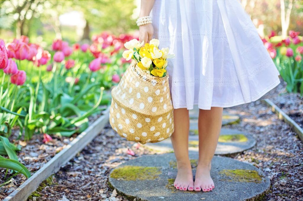 Detail na bosé nohy dívky v bílých šatech s taškou plnou jarních květin.