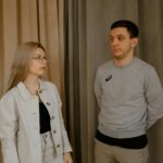 A young couple stands facing each other, appearing to be in a thoughtful discussion indoors.